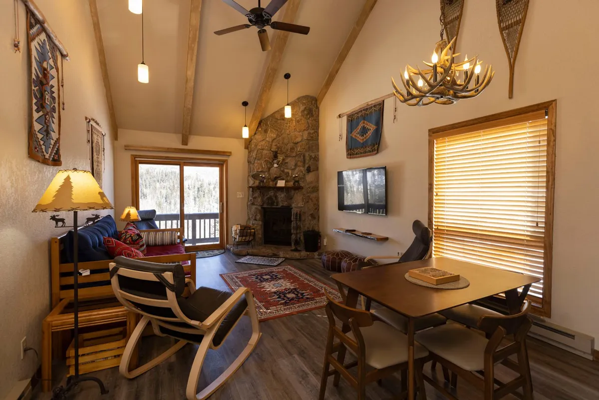 This cozy interior scene features a rustic stone fireplace with a wooden mantel, a classic rocking chair, colorful patterned rugs, and a wood dining table—evoking a sense of warmth and relaxation. Natural light streams in, hinting at mountain views from the balcony, and the inviting furnishings create a perfect space for unwinding after outdoor adventures.