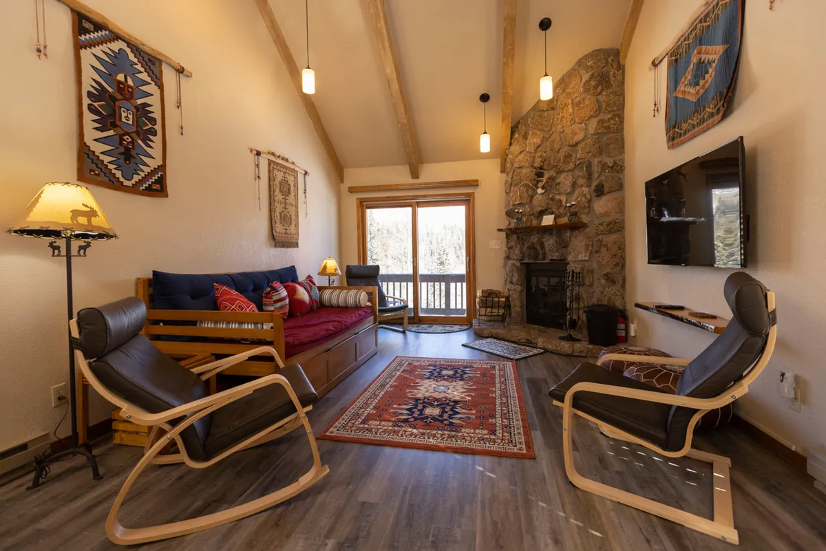 A comforting living area with a stone fireplace, a colorful patterned rug, a wooden futon with plush cushions, and two modern rocking chairs. The space is arranged for relaxed conversation or reading, with a view through the balcony door hinting at the alpine setting.
