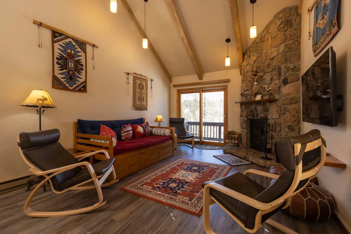 A cozy interior space featuring a stone fireplace, wooden daybed with colorful cushions, rocking chairs, and woven tribal-patterned rugs. The seating area is warm and inviting, perfect for relaxing after a day of outdoor activities. Large windows or sliding doors suggest a connection to the mountain landscape outside.