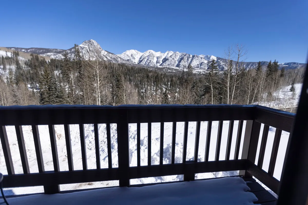 An exterior balcony scene with a snow-covered wooden railing, distant snow-capped mountains, and bare winter trees under a vibrant blue sky. The view is expansive, emphasizing the grandeur of the alpine environment and the opportunity to relax outdoors amid serene winter surroundings.