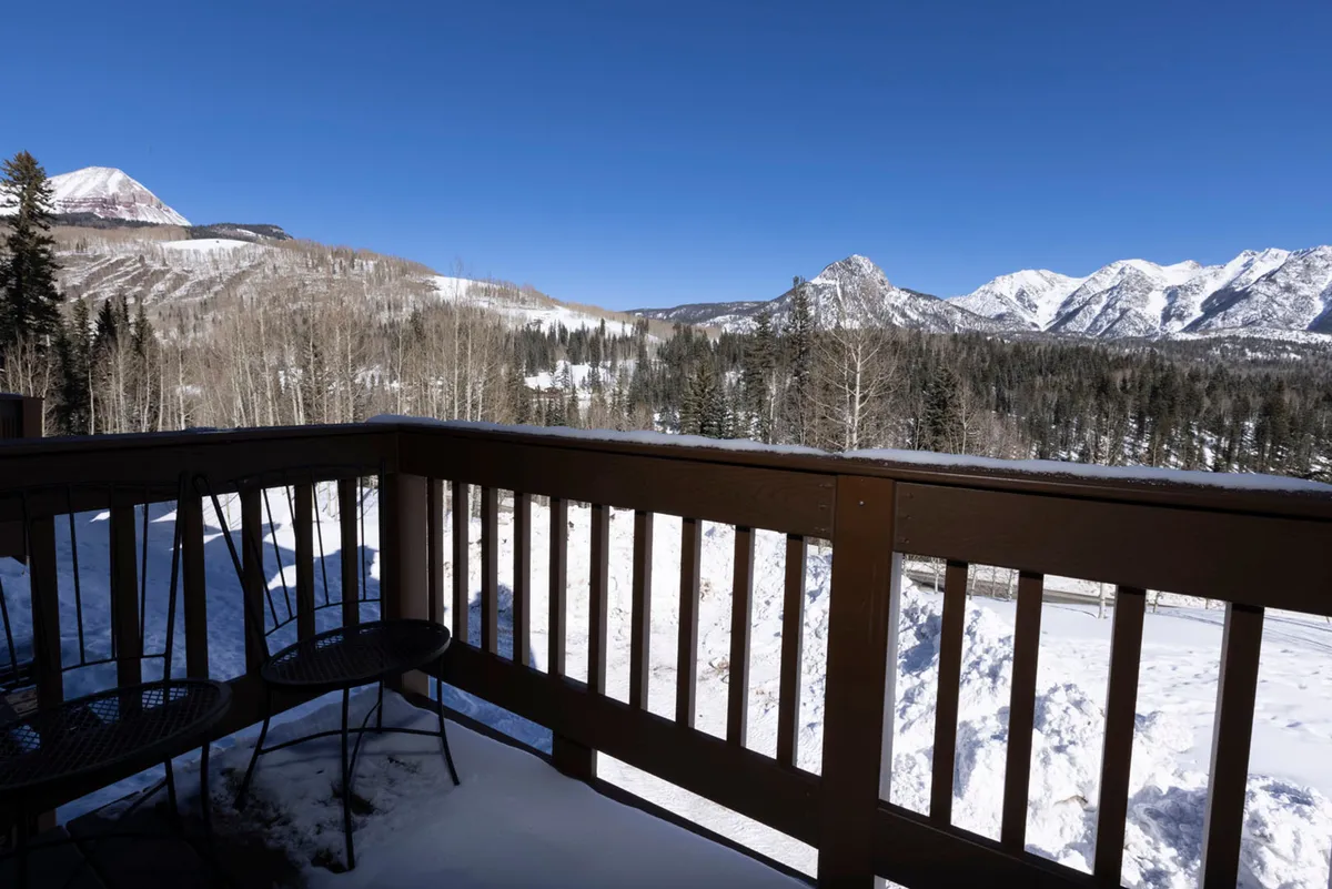 A peaceful wooden balcony overlooking a breathtaking panorama of snow-covered mountain peaks. Bare aspen trees frame the view, while metal chairs dusted with snow invite guests to sit and soak in the tranquil winter landscape. The setting radiates privacy and a deep connection to nature, evoking the feeling of a secluded mountain retreat.