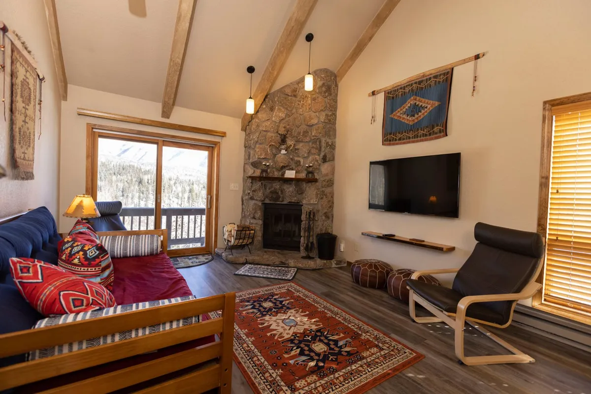This image features a stone fireplace with a mantle as the focal point, surrounded by colorful patterned cushions and a rug, and a large window that frames a breathtaking mountain view. A wall-mounted flat screen TV adds a touch of modern comfort, while the seating area invites relaxation by the fire after a day outdoors.