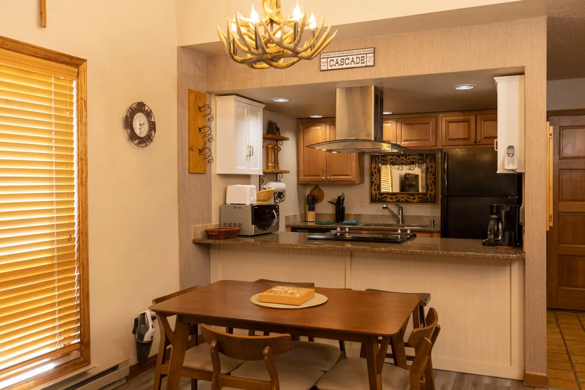 A wooden dining table with a centerpiece sits adjacent to a kitchen area featuring a granite countertop and modern appliances. An antler chandelier and a decorative 'Cascade' sign above the kitchen pass-through add mountain lodge character. The space feels inviting and well-lit, ideal for meal preparation and casual gatherings.