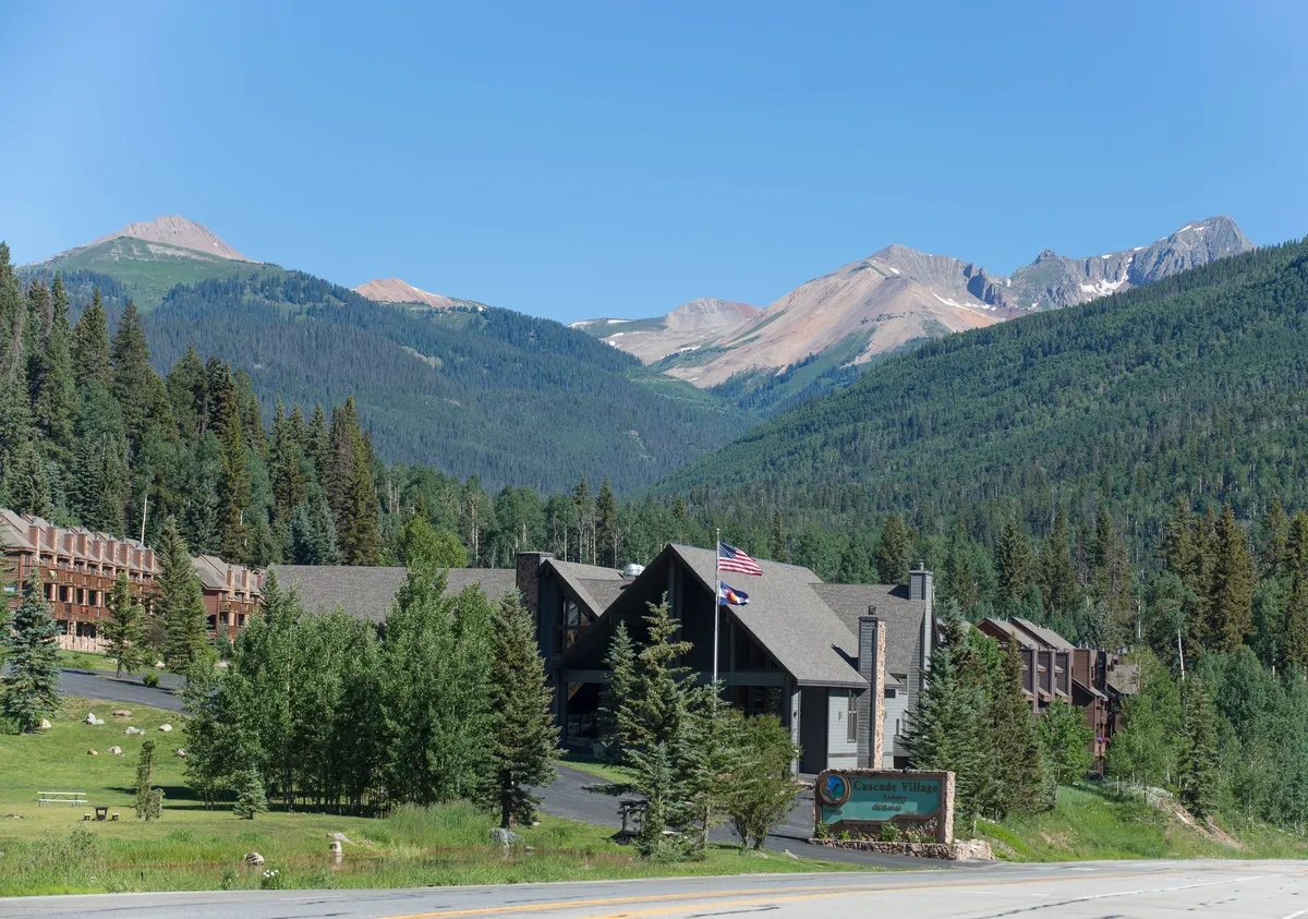 A welcoming exterior shot of the Cascade Village lodge building with its peaked roof and stone chimney, surrounded by dense evergreen trees and landscaped areas. The prominent signage at the entrance and distant mountain peaks set the scene for a mountain adventure base. The image subtly hints at amenities such as the ski shuttle and ski storage, integral to the lodge experience.