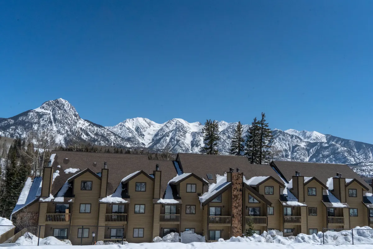 A multi-unit chalet-style building with balconies is nestled among tall evergreen trees, all blanketed in snow. Snow covers the rooftops and grounds, and majestic mountain peaks rise in the background. The scene is peaceful yet invigorating, suggesting both proximity to mountain activities and the comforts of a mountain retreat.