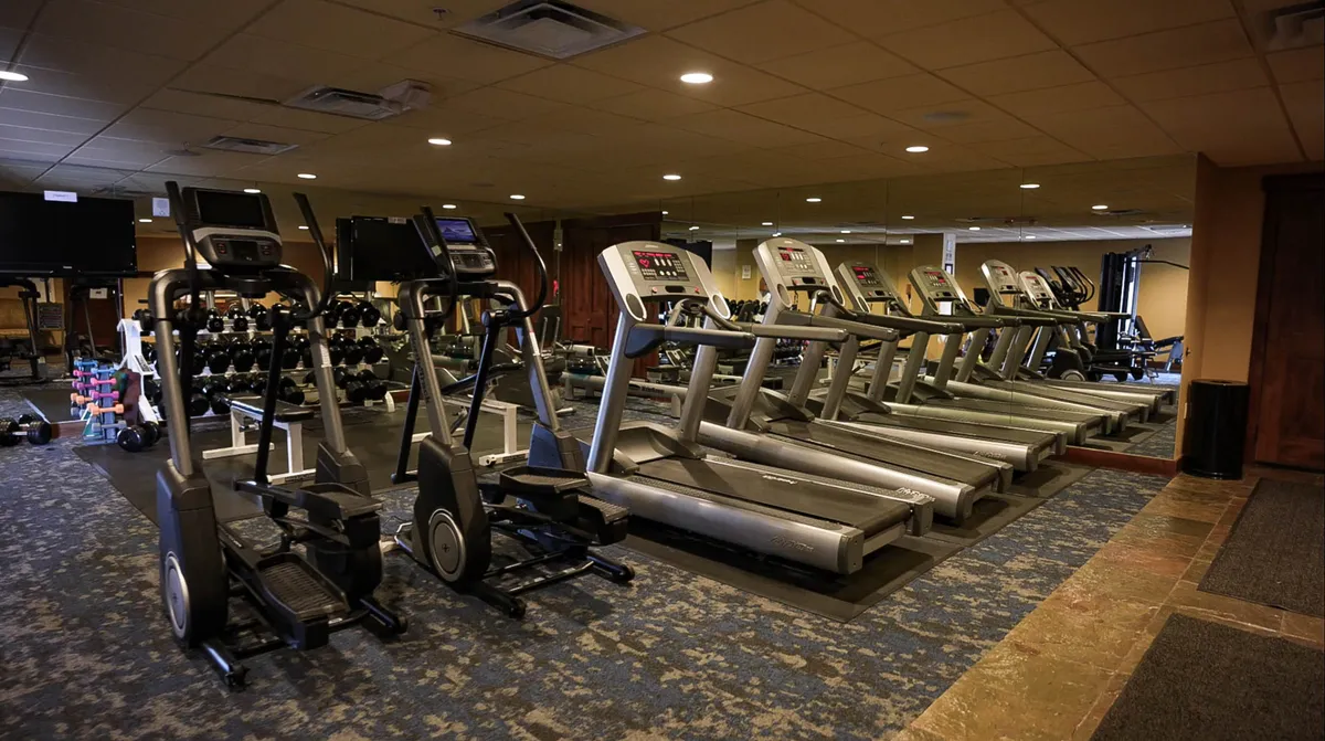 This image showcases a well-equipped fitness center with a row of treadmills, elliptical machines, a dumbbell rack, and exercise mats. The space is brightly lit and designed for a variety of workouts, from cardio to strength training.