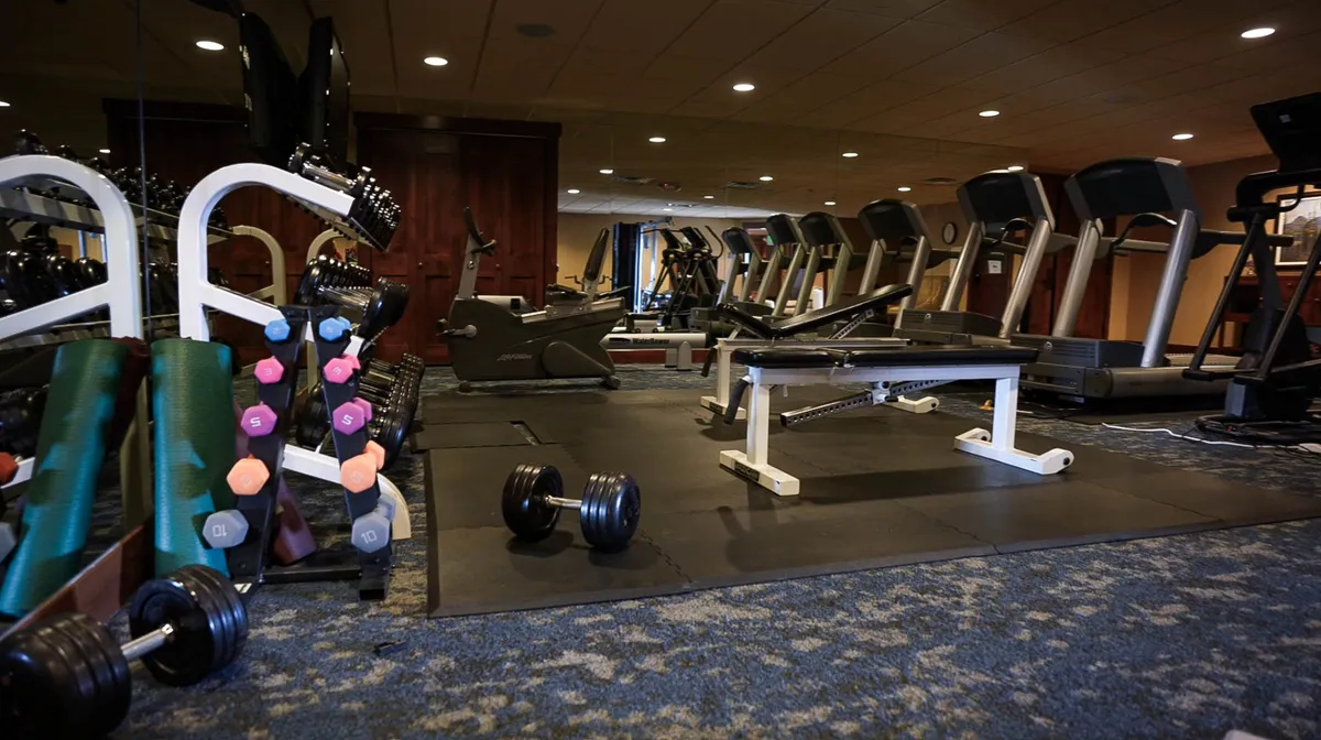 A spacious and motivating fitness center featuring a row of treadmills along the back wall, a dumbbell rack with colorful weights, a weight bench, and various cardio machines including stationary bikes. The environment encourages both solo and group fitness activities.