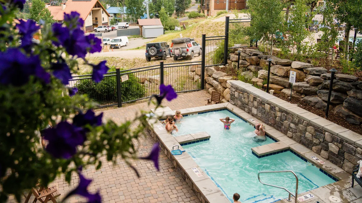 An inviting heated outdoor pool is surrounded by a stone wall and a paved patio with chairs, with people enjoying the water. Purple flowers add a touch of color to the foreground, and the mood is leisurely and social.