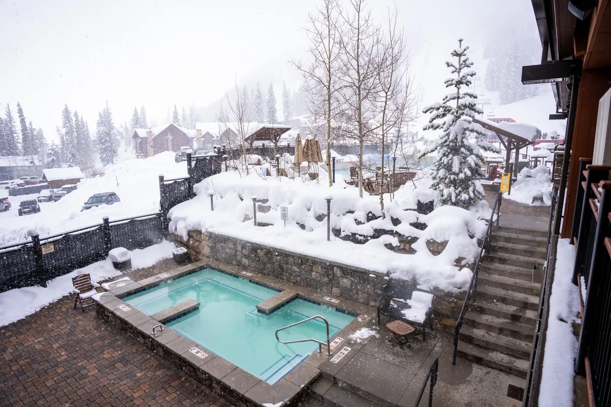 This image features a heated outdoor pool with bright turquoise water, surrounded by snow-covered stone walls and patio. Stairs lead up to an outdoor seating area, and snow-laden trees and umbrellas accentuate the tranquil winter setting. The inviting pool and nearby hot tub are the clear focus, with a backdrop of serene mountain scenery.