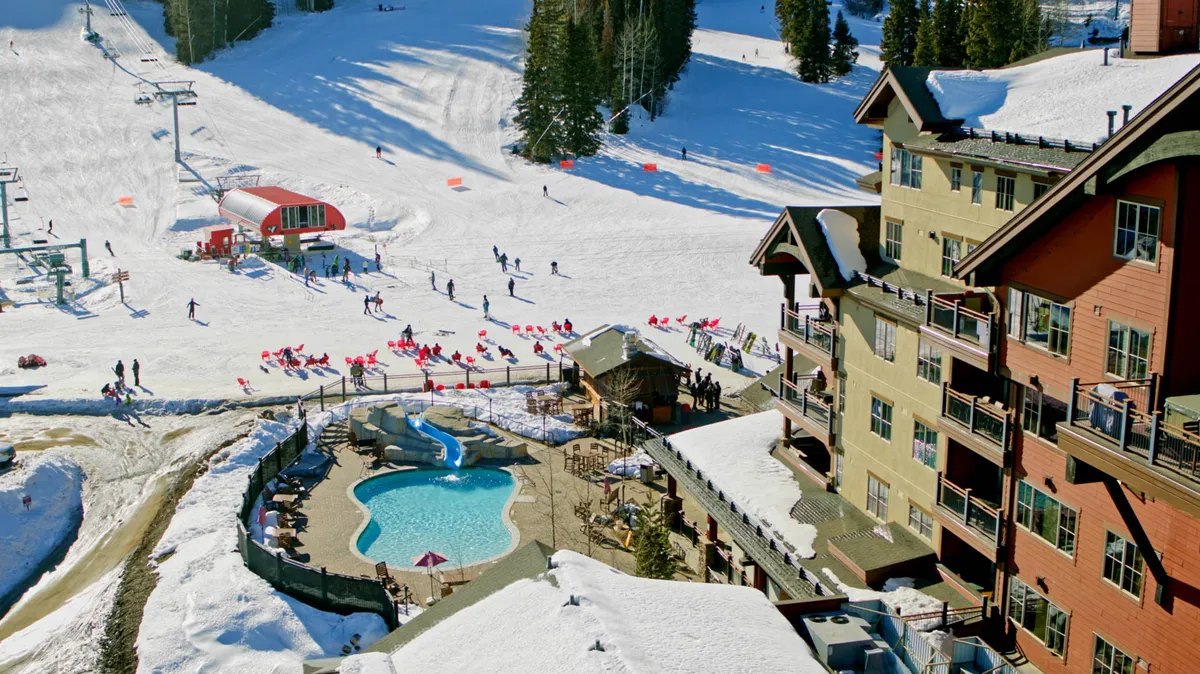 This lively exterior shot captures the heated outdoor pool and hot tub adjacent to the resort building, framed by snow, with skiers and snowboarders visible on the nearby slopes. Red chairs and balconies offer spots to relax and enjoy panoramic mountain views, emphasizing the blend of recreation and relaxation.