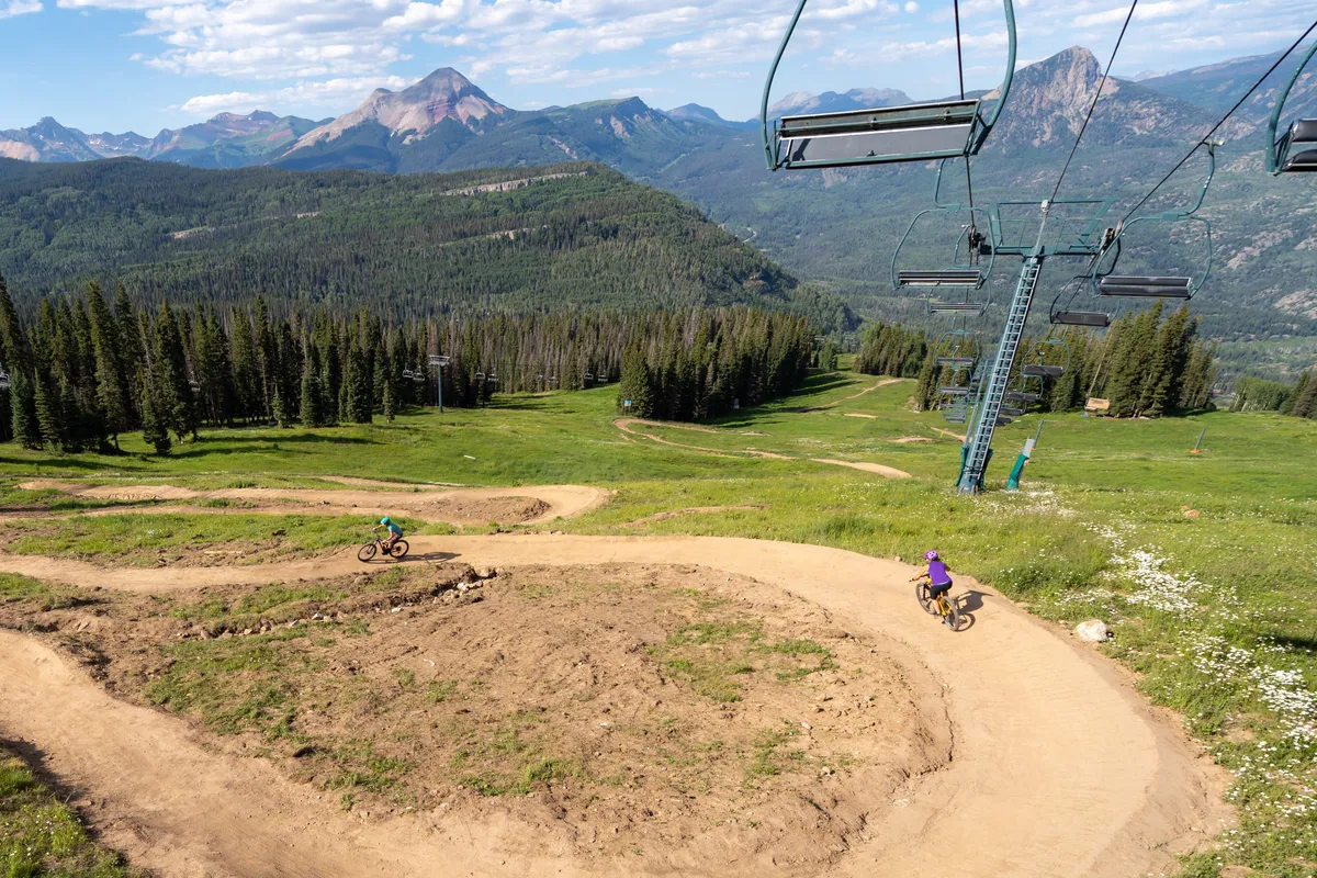 An energizing exterior view of a mountain bike trail winding down a hillside, with two cyclists riding along the path. A ski lift chair is visible above, framed by distant mountain peaks and forested terrain. This dynamic scene showcases both winter and summer adventure possibilities, with infrastructure (like the ski lift) that hints at convenient access and storage for gear.