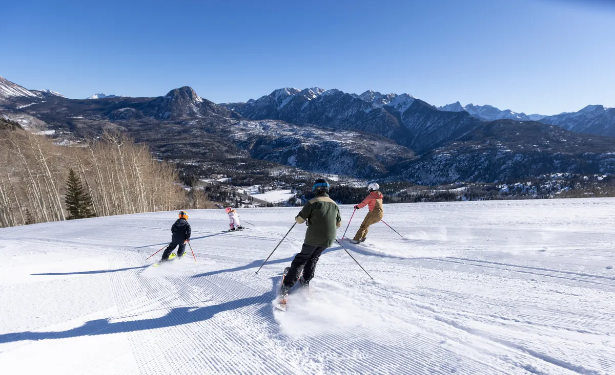This exterior shot shows four skiers gliding down a pristine, groomed slope with a breathtaking snow-capped mountain range in the background. The clear blue sky and leafless trees at the edge of the slope add to the invigorating, active winter atmosphere. The image captures the thrill and energy of being on the mountain, surrounded by natural beauty.